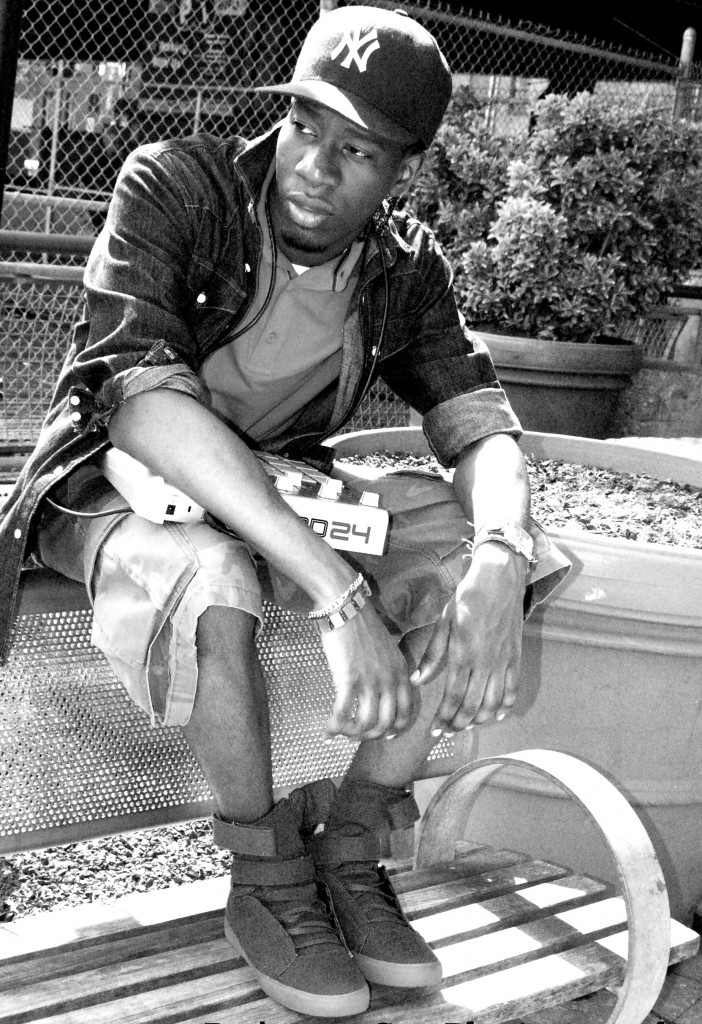 Headshot of Producer's Toolbox artist, Samuel Lee aka SammayOnTheBeat. He sits on top of the back of a park bench with a synth in his lap, a baseball cap, shorts, jean jacket, polo, sneakers, staring off into the distance.
