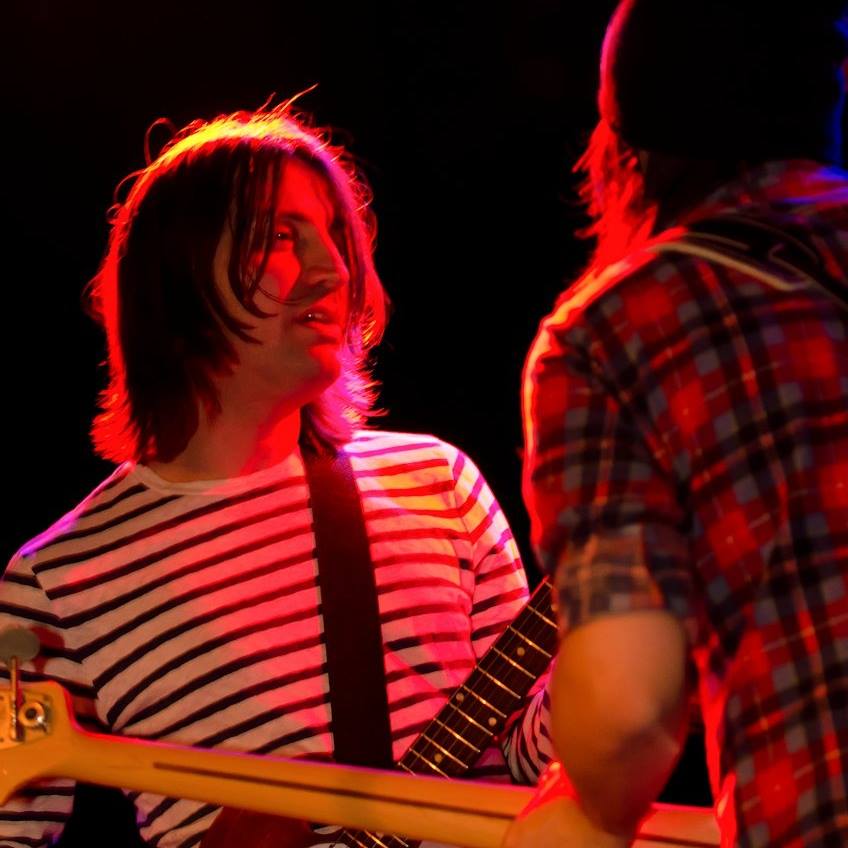 Josh Lattanzi in a black and white striped shirt playing guitar and looking at his bandmate on stage. Josh Lattanzi is a composer with Flavorlab's exclusive music catalog, Producer's Toolbox.