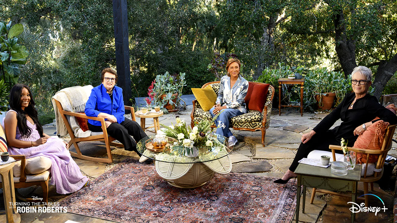 Shot from Turning The Tables with Robin Roberts. Robin and her guests are sitting outside on a patio in four cushioned chairs around an oriental rug and wicker center table made to look like a flower with a round glass table top. From the left Mickey Guyton in a lavender dress, Billie Jean King in a royal blue blazer and black slacks, Robin Roberts in a flower pale blue top and blue pants, and Jamie Lee Curtis in a black jumpsuit are turning to smile at the camera. Flavorlab Sound provided audio post and Flavorlab Score provided original music to the series.